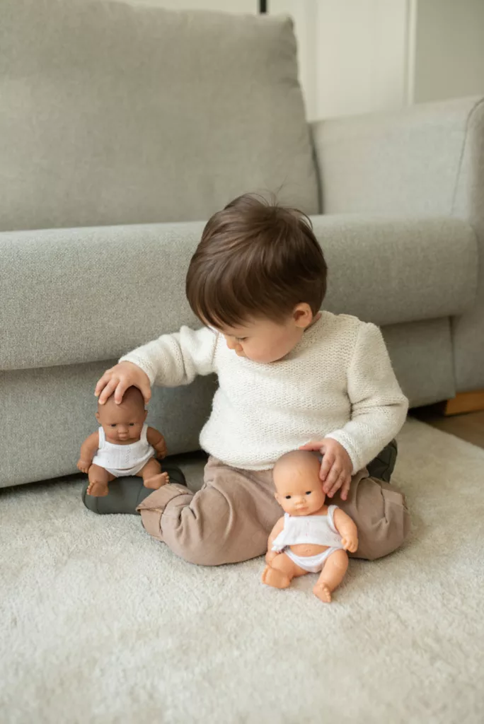 A young child sits on a carpeted floor in front of a gray couch, holding two baby dolls. The child is wearing a cream-colored sweater and beige pants. One doll with darker skin is seated on a small green cushion, while the child gently holds the head. The other doll with lighter skin rests on the child’s lap.