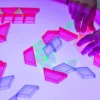 Children's hands arranging colorful translucent geometric shapes on a glowing light table. The shapes include pink trapezoids, green triangles, and blue rectangles, which are illuminated and glowing vividly under the light.