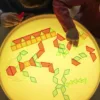 Children's hands arranging colorful translucent geometric shapes on a glowing light table. The shapes include pink trapezoids, green triangles, and blue rectangles, which are illuminated and glowing vividly under the light.
