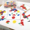 A tabletop setup with colorful pattern blocks and activity cards used for shape and design learning. A clear plastic container holds various geometric pieces in red, yellow, green, blue, and orange. Several activity cards are spread out, some partially completed with pattern blocks placed on outlined designs, showing imaginative shapes and objects. The scene encourages hands-on play and creativity in a classroom or educational setting.