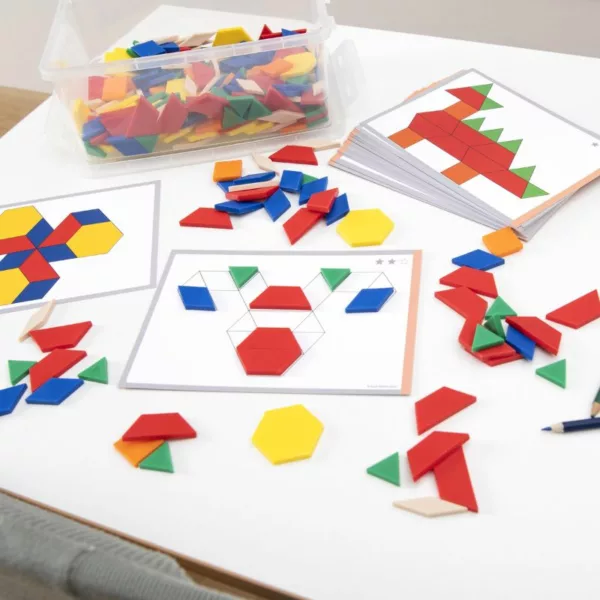 A tabletop setup with colorful pattern blocks and activity cards used for shape and design learning. A clear plastic container holds various geometric pieces in red, yellow, green, blue, and orange. Several activity cards are spread out, some partially completed with pattern blocks placed on outlined designs, showing imaginative shapes and objects. The scene encourages hands-on play and creativity in a classroom or educational setting.