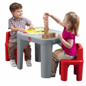 A young boy and a young girl are sitting at a gray plastic table with red chairs, playing with wooden blocks. The girl is stacking the blocks, which have numbers and math symbols on them, while the boy watches her and smiles.