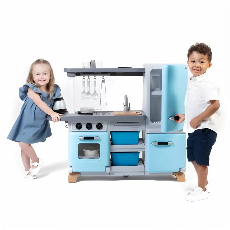 A young girl and a young boy stand on opposite sides of a blue and gray Step2 Cooking Time Kitchen playset, smiling at the camera. The kitchen has a stove, oven, sink, and refrigerator.