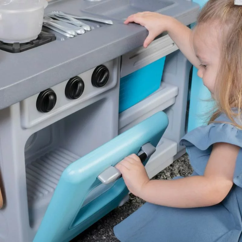 A young child with blonde hair, wearing a blue dress, kneels on the floor and opens the oven door of a gray and blue toy kitchen set. The toy kitchen has a stovetop with a pot and some utensils.