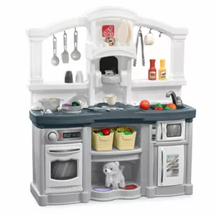 A gray and white Step2 Fun with Friends toy kitchen playset with a pretend oven, microwave, sink, and coffee maker. The playset is filled with toy food, pots, pans, and utensils. A small stuffed dog toy is placed on a shelf below the counter.