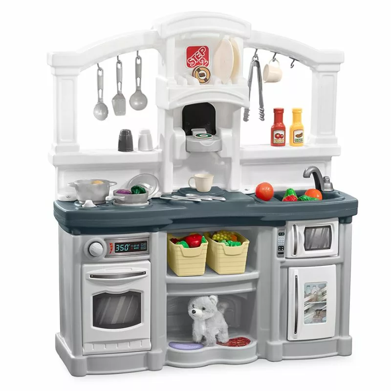 A gray and white Step2 Fun with Friends toy kitchen playset with a pretend oven, microwave, sink, and coffee maker. The playset is filled with toy food, pots, pans, and utensils. A small stuffed dog toy is placed on a shelf below the counter.