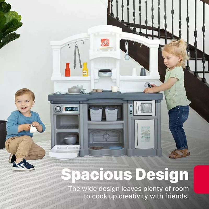 A young child with blonde hair, wearing a blue dress, kneels on the floor and opens the oven door of a gray and blue toy kitchen set. The toy kitchen has a stovetop with a pot and some utensils.