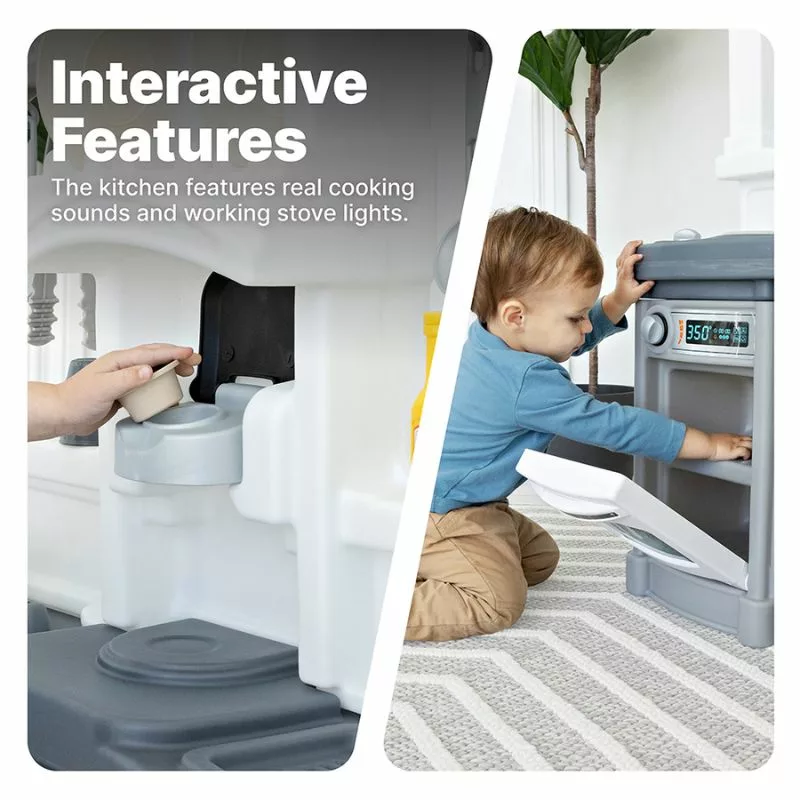 A young child with blonde hair, wearing a blue dress, kneels on the floor and opens the oven door of a gray and blue toy kitchen set. The toy kitchen has a stovetop with a pot and some utensils.