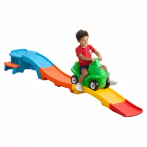A young child in a red shirt and shorts smiles while riding a green toy roller coaster car down a colorful plastic track. The track is made of blue, orange, yellow, and red sections.