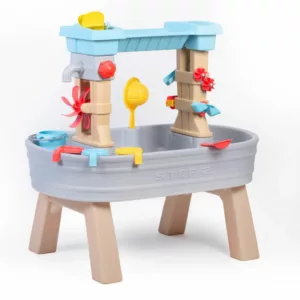 A Step2 Rain Showers & Flow Water Table, a gray and blue plastic toy with a raised water channel, a red water wheel, and various yellow and red scoops and toys in the basin. The text "STEP 2" is visible on the side of the table.