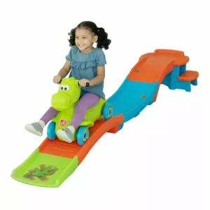 A young girl with pigtails smiles as she rides a green dinosaur-themed Step2 Dino Dash Up & Down Roller Coaster toy on a multi-colored track.