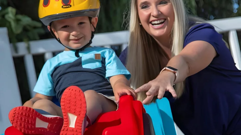 A smiling woman pushes a child in a yellow flame helmet down a red and blue toy roller coaster ramp outdoors.