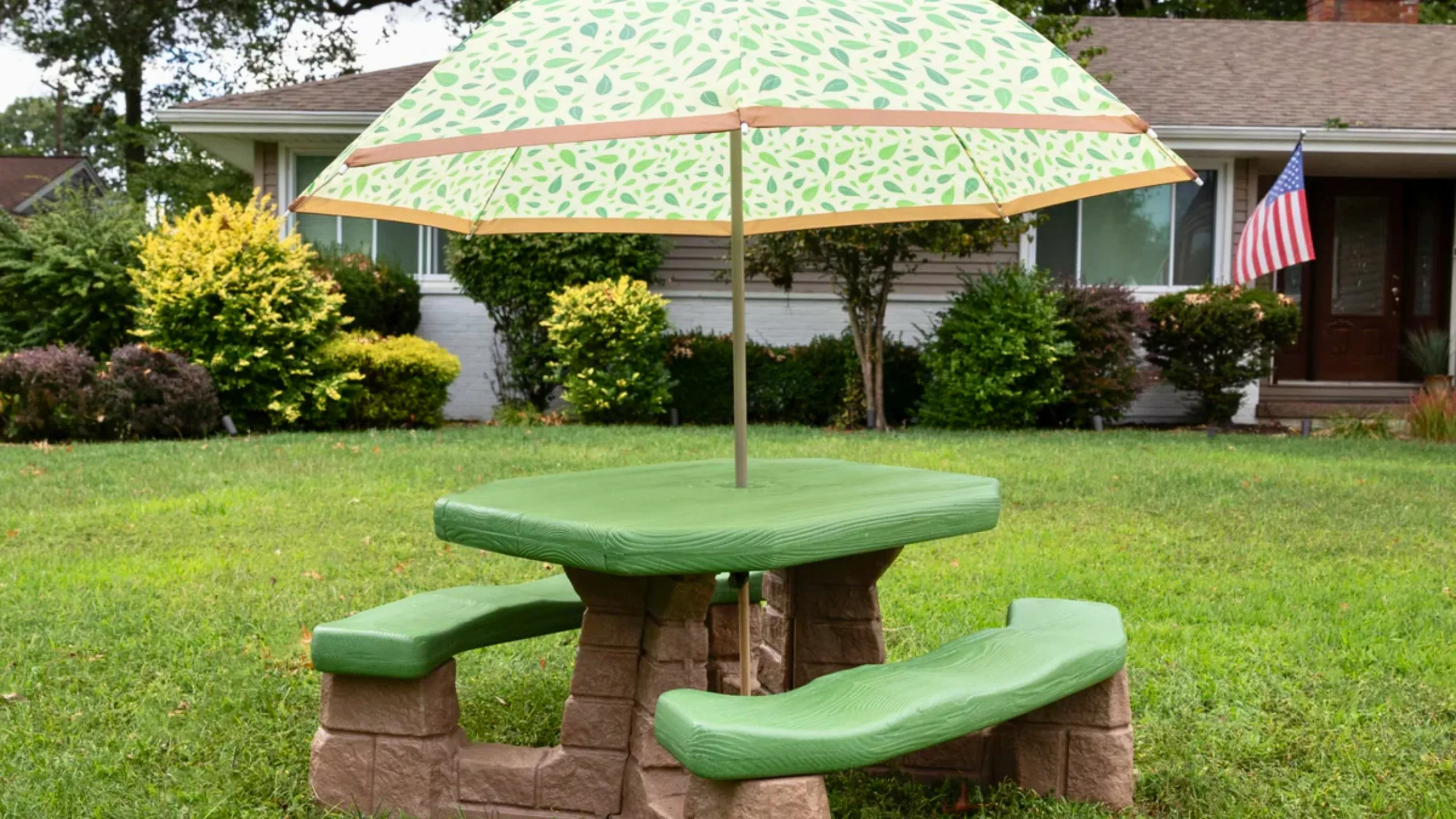 A green and tan Step2 kids' picnic table with a leaf-patterned umbrella sits on a grassy lawn in front of a house.